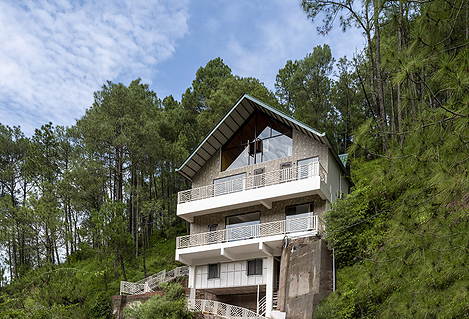 Modern hillside house with multiple balconies surrounded by tall pine trees and lush greenery.